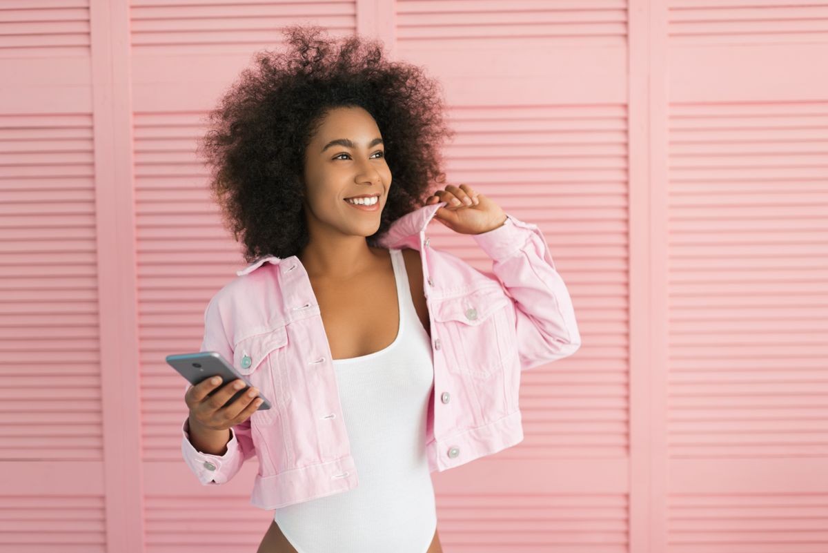 Positive African American woman with beautiful smile and sexy body holding mobile phone standing near pink background. Happy curly hair hipster wearing stylish jeans jacket in showroom. Shopping.